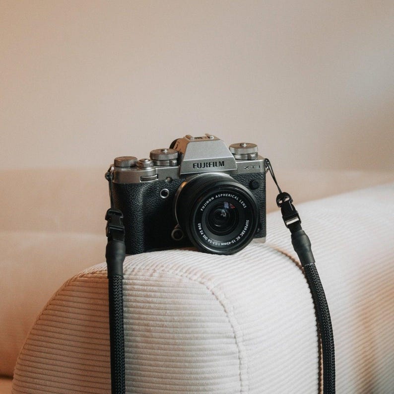 Pu&ograve; includere: Una fotocamera digitale mirrorless Fujifilm X-T1 con corpo nero e argento e obiettivo nero. La fotocamera ha un cinturino nero attaccato. La fotocamera &egrave; appoggiata su una superficie chiara e strutturata.