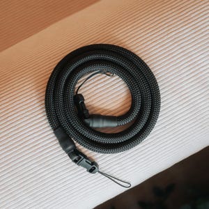 Pu&ograve; includere: Una tracolla nera per fotocamera, arrotolata su una superficie. La tracolla ha una trama intrecciata e una fibbia di plastica. Include un piccolo anello per il fissaggio a una fotocamera.