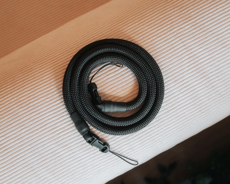 Pu&ograve; includere: Una tracolla nera per fotocamera, arrotolata su una superficie. La tracolla ha una trama intrecciata e una fibbia di plastica. Include un piccolo anello per il fissaggio a una fotocamera.