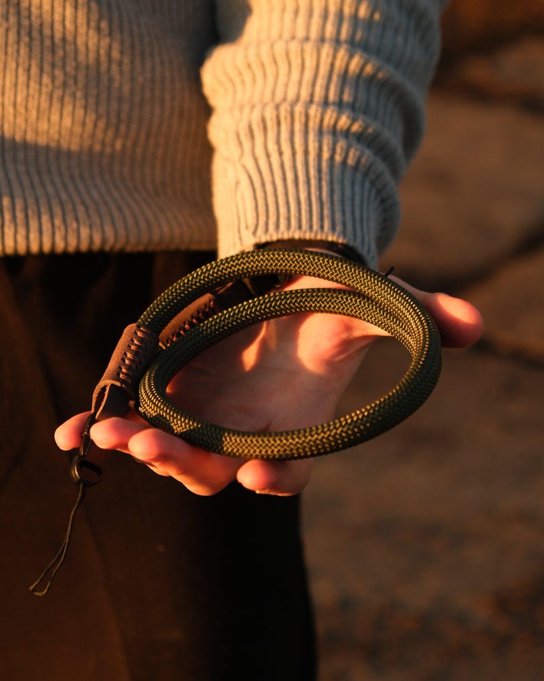 May include: A coiled olive green rope wrist strap with a brown leather accent and a black clasp. The rope has a textured, braided appearance. The strap is held in a hand, with a neutral background.