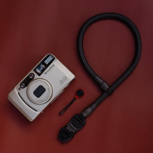 Pode incluir: Uma câmara compacta prateada Fujifilm DL-270 Zoom Super com uma lente Fujinon Zoom 38-70mm, acompanhada por uma correia de corda entrançada preta com uma fivela de plástico de libertação rápida e um pequeno acessório circular vermelho e preto.