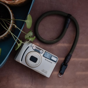 Pode incluir: Câmera Fujifilm DL-270 Zoom Super prateada com lente e flash pretos. Possui uma alça tecida em verde escuro e preto. A câmera está em uma superfície marrom com uma planta ao fundo.