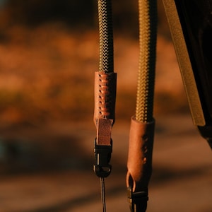 May include: Close-up of two olive green camera straps with brown leather accents. The straps have a woven texture and are attached to black plastic buckles. The background is blurred, with warm tones.