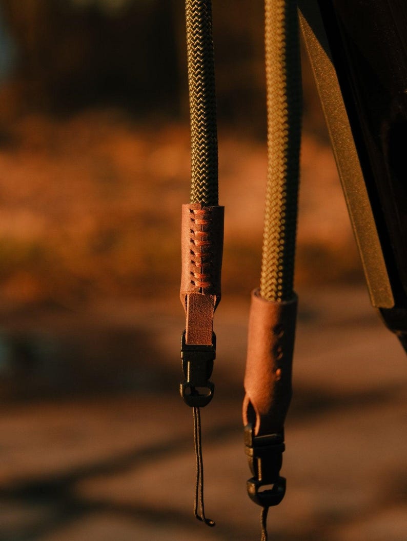 May include: Close-up of two olive green camera straps with brown leather accents. The straps have a woven texture and are attached to black plastic buckles. The background is blurred, with warm tones.