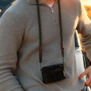May include: A black digital camera with a lens is suspended by a green and brown camera strap. The camera hangs against a light gray sweater with a zipper. The camera strap is attached to the camera with black clips.