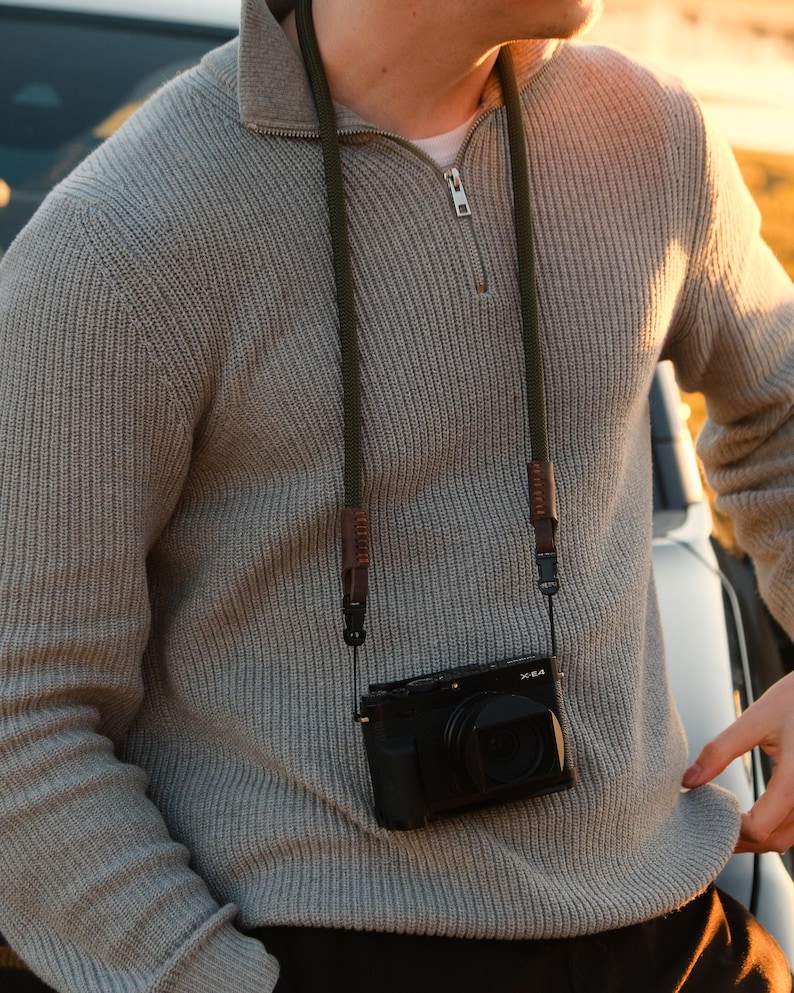 May include: A black digital camera with a lens is suspended by a green and brown camera strap. The camera hangs against a light gray sweater with a zipper. The camera strap is attached to the camera with black clips.
