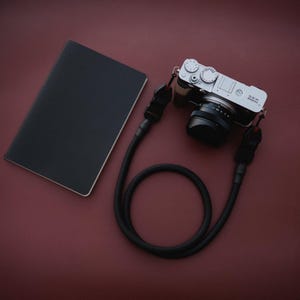 May include: A silver digital camera with a black lens and a black camera strap lies next to a black notebook on a maroon surface. The camera strap is looped.