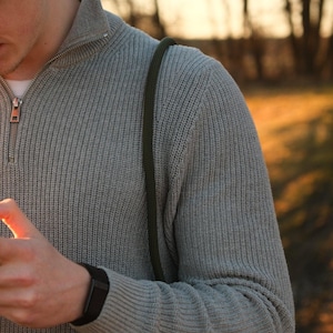 May include: A light gray knit sweater with a quarter-zip collar. The sweater has a ribbed texture and a black watch on the wrist. A green strap is visible over the shoulder. The background is blurred with trees and sunlight.