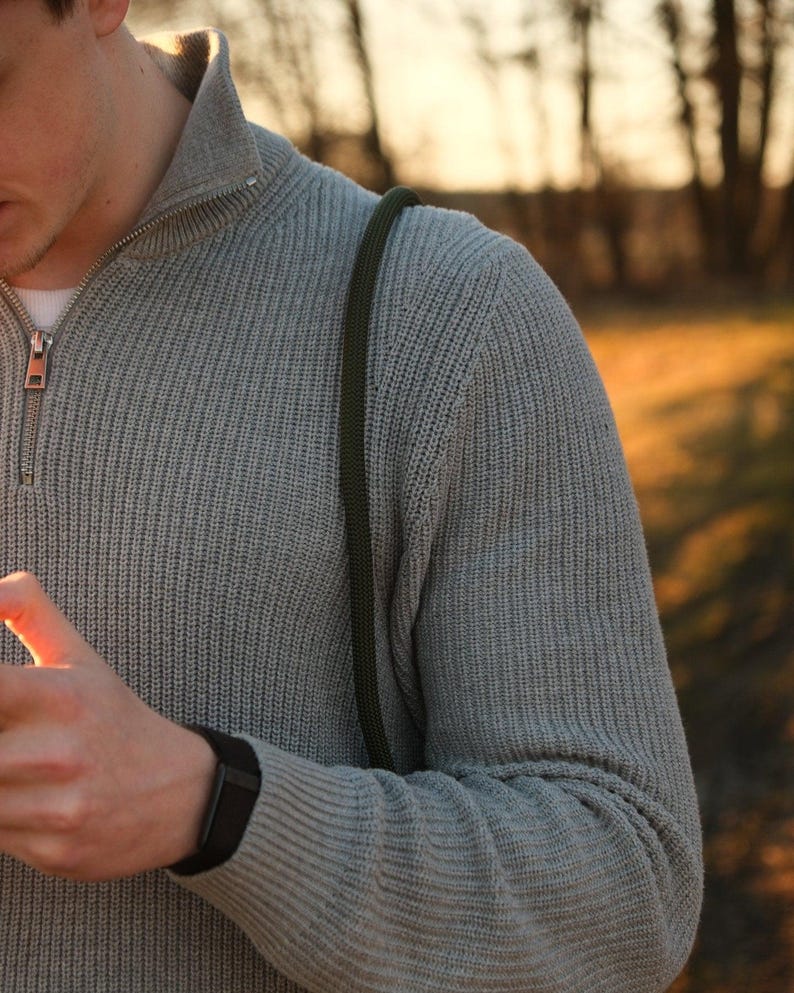 May include: A light gray knit sweater with a quarter-zip collar. The sweater has a ribbed texture and a black watch on the wrist. A green strap is visible over the shoulder. The background is blurred with trees and sunlight.