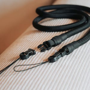 Pu&ograve; includere: Un cordino nero intrecciato con un fermaglio in plastica e un piccolo anello. Il cordino &egrave; progettato per essere indossato al collo e ha un aspetto strutturato. Il fermaglio &egrave; aperto e il cordino &egrave; appoggiato su una superficie chiara.