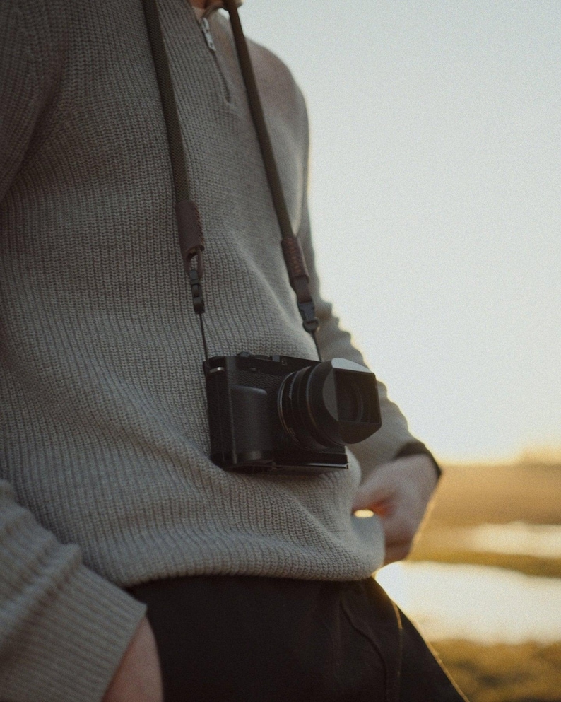 Könnte beinhalten: Eine schwarze Digitalkamera mit Objektiv hängt an einem dunkelbraunen Riemen um den Hals. Die Kamera hängt an einem hellgrauen Pullover. Der Hintergrund zeigt eine verschwommene Feld- und Himmelsansicht.