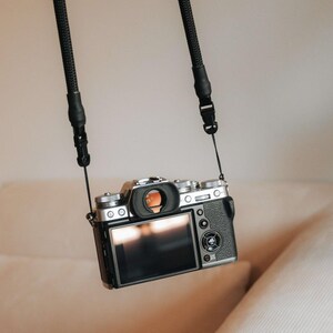 Pu&ograve; includere: Una fotocamera digitale nera e argento &egrave; sospesa da una cinghia nera. La fotocamera ha un ampio schermo LCD e diverse manopole e pulsanti. La cinghia &egrave; realizzata in materiale intrecciato ed &egrave; attaccata alla fotocamera in due punti.