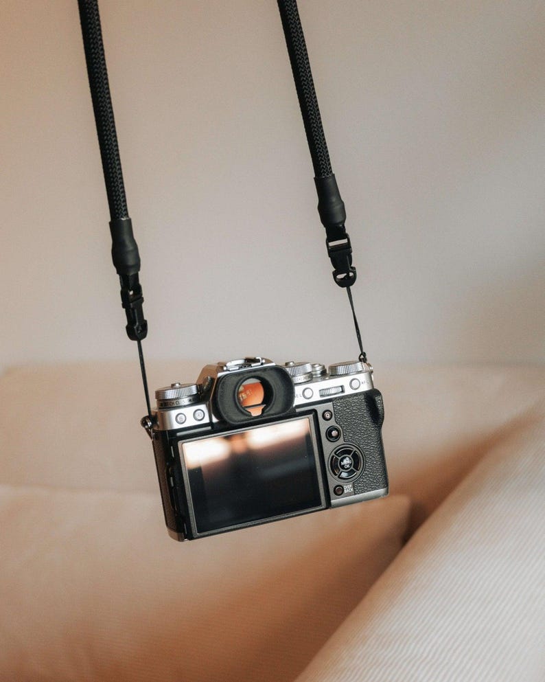 Pu&ograve; includere: Una fotocamera digitale nera e argento &egrave; sospesa da una cinghia nera. La fotocamera ha un ampio schermo LCD e diverse manopole e pulsanti. La cinghia &egrave; realizzata in materiale intrecciato ed &egrave; attaccata alla fotocamera in due punti.