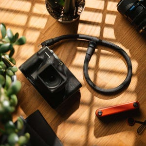May include: A black digital camera, a black camera strap, and a red and black multi-tool are arranged on a wooden surface. The camera lens is facing forward. The scene is lit by natural light.