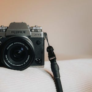 Pu&ograve; includere: Una fotocamera Fujifilm X-T3 argento e nera con obiettivo e cinturino neri. La fotocamera &egrave; su una superficie bianca. L'obiettivo ha la scritta "FUJINON ASPHERICAL LENS" e il corpo macchina "FUJIFILM X-T3".