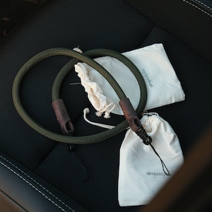 May include: Two olive green camera straps with brown leather accents and black clasps are displayed. Two small off-white drawstring bags and a larger off-white bag with the text "strapstyle" are also present. The items are arranged on a black surface.