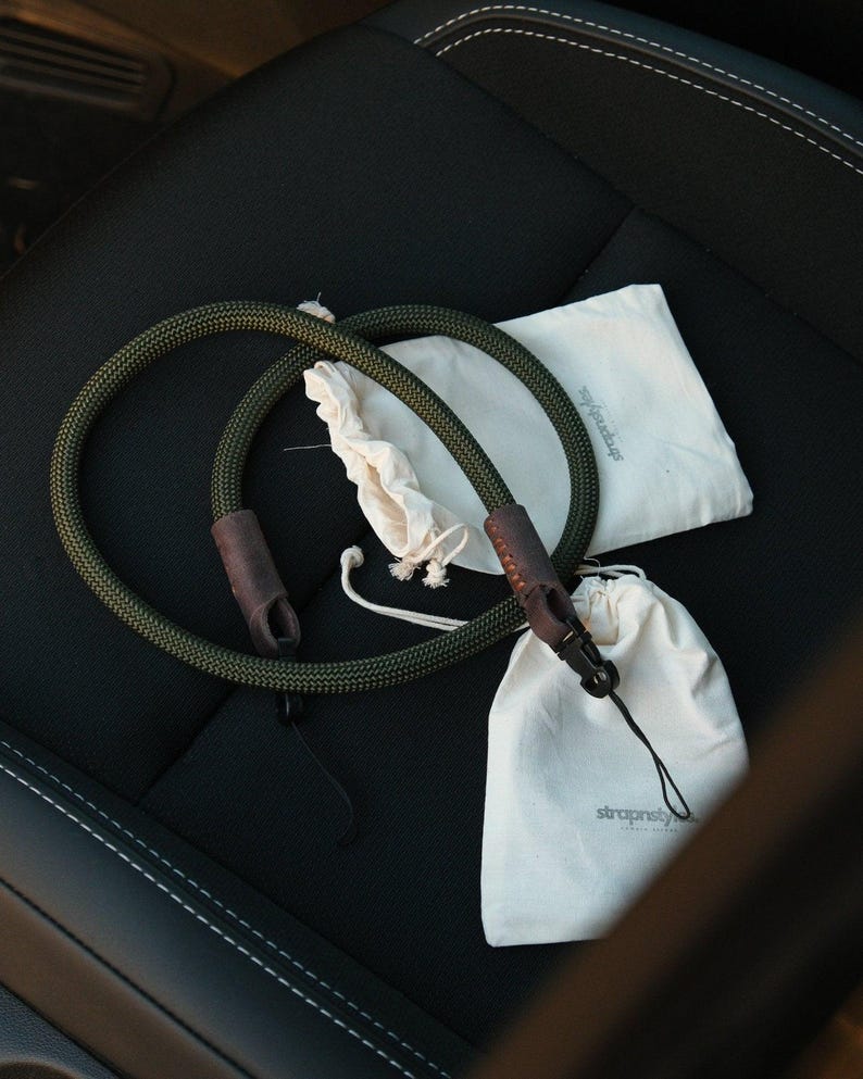 May include: Two olive green camera straps with brown leather accents and black clasps are displayed. Two small off-white drawstring bags and a larger off-white bag with the text "strapstyle" are also present. The items are arranged on a black surface.