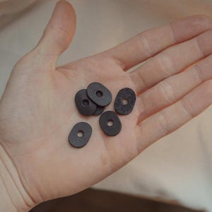 Könnte beinhalten: Eine Hand hält mehrere kleine, dunkelgraue, ovale Unterlegscheiben mit einem Loch in der Mitte. Eine Unterlegscheibe hat eine strukturierte Oberfläche. Die Unterlegscheiben bestehen wahrscheinlich aus einem haltbaren Material und sind für verschiedene Anwendungen geeignet.