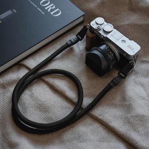 Pode incluir: Uma câmara digital Fujifilm X-E3 prateada com lente preta e punho de madeira castanha, presa a uma correia de corda entrançada preta e espessa. A câmara possui um sistema de seletores no topo e está posicionada ao lado de um livro preto.