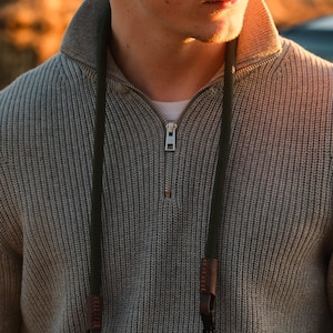 May include: A gray knit sweater with a quarter-zip collar and a dark green camera strap. The camera strap has brown leather accents and is attached to the sweater. The sweater is a neutral color and has a ribbed texture.