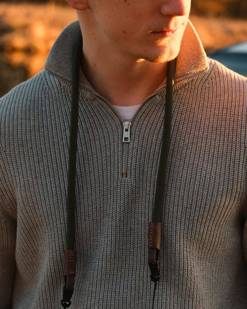 May include: A gray knit sweater with a quarter-zip collar and a dark green camera strap. The camera strap has brown leather accents and is attached to the sweater. The sweater is a neutral color and has a ribbed texture.