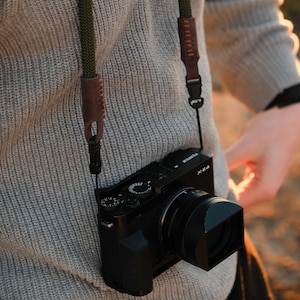 May include: Black digital camera with a lens hood, hanging from a green and brown camera strap. The camera is suspended against a gray sweater. The strap has leather accents and is attached to the camera.
