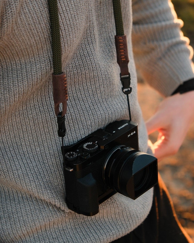 May include: Black digital camera with a lens hood, hanging from a green and brown camera strap. The camera is suspended against a gray sweater. The strap has leather accents and is attached to the camera.