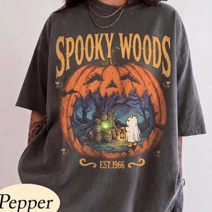 May include: A charcoal gray t-shirt featuring a large orange pumpkin graphic with the text SPOOKY WOODS at the top and EST. 1966 at the bottom. Inside the pumpkin is a forest scene with a bear in a ghost costume holding a lantern near honey pots.