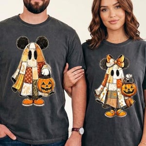 May include: Two charcoal gray t-shirts featuring a graphic of a ghost character dressed in a patchwork quilt pattern. The left shirt displays a ghost with round ears, while the right shirt shows a ghost with a bow and ears, both holding a pumpkin bucket.
