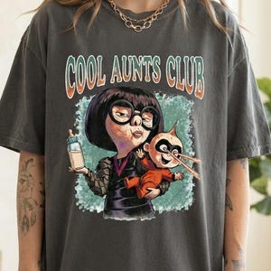 Puede incluir: Una camiseta gris carbón de manga corta con el texto COOL AUNTS CLUB en una fuente retro. El gráfico muestra a un personaje de dibujos animados con pelo negro y gafas sosteniendo un biberón, mientras carga a un bebé con un traje de superhéroe rojo.