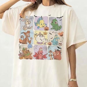 Puede incluir: Una camiseta de color marfil de manga corta con un estampado gráfico en cuadrícula de varios personajes animados, incluyendo un conejo, pez, lagarto, mono, tetera, criatura azul, oso, cangrejo y facóquero, decorado con calabazas y murciélagos.