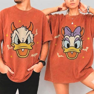 以下が含まれることがあります： 錆色の半袖Tシャツ2枚。漫画のキャラクターのダックがプリントされています。左のTシャツにはオレンジ色の角が生えたダック、右のTシャツには紫色のリボンをつけたダックが描かれています。どちらのシャツにも小さな黄色い星とコウモリの模様があしらわれています。