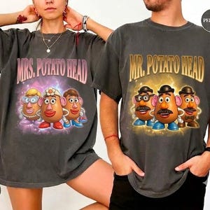 May include: Two charcoal gray t-shirts featuring graphic prints. The left shirt displays three Mrs. Potato Head characters with the text MRS. POTATO HEAD. The right shirt displays three Mr. Potato Head characters with the text MR. POTATO HEAD.