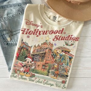 Peut inclure: Un t-shirt couleur crème avec un imprimé de Disney's Hollywood Studios, incluant le Hollywood Tower Hotel, Mickey Mouse en bonnet de Père Noël et Pluto. Le texte indique Disney's Hollywood Studios et That Never Was And Always Will Be.