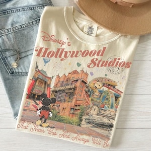 Może przedstawiać: Kremowy t-shirt z nadrukiem graficznym przedstawiającym Disney's Hollywood Studios z Myszką Miki, hotelem Hollywood Tower i Slinky Dog Dash. Tekst głosi Disney's Hollywood Studios oraz That Never Was And Always Will Be.