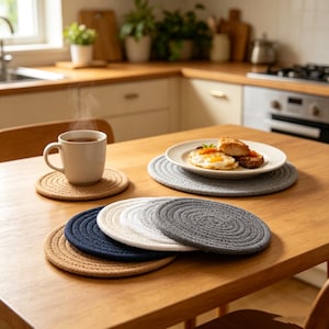 May include: A collection of round, woven rope coasters and placemats in neutral and dark tones including beige, navy blue, white, and grey. The items feature a spiral pattern and are arranged on a wooden table alongside a steaming mug and a plate.