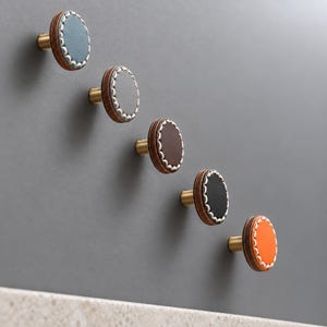 May include: Five circular cabinet knobs arranged diagonally. Each knob features a central colored disc in blue, gray, brown, black, or orange, surrounded by a white stitched border and a dark wood-toned edge, mounted on a gold-toned metal base.