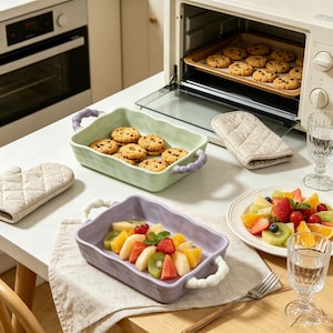 May include: A light green rectangular ceramic baking dish with purple beaded handles containing chocolate chip cookies, and a light purple rectangular ceramic baking dish with white beaded handles containing a fresh fruit salad.