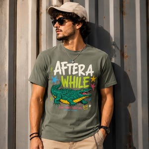 Peut inclure: T-shirt vert olive avec un motif coloré d'un alligator vert et le texte "AFTER A WHILE". Le design comprend des étoiles et la phrase "GOOD THINGS TAKE TIME". Le t-shirt est porté par une personne portant des lunettes de soleil et une casquette.