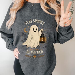 Könnte beinhalten: Ein dunkelgraues Sweatshirt mit Rundhalsausschnitt und einem Grafikdruck eines lächelnden Geistes, der eine leuchtende Laterne hält. Der Text STAY SPOOKY steht über dem Geist und BE KIND BE WICKED darunter, ergänzt durch ein Kürbis-Symbol.