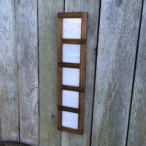 May include: A vertical wooden picture frame with four white square openings. The frame is made of dark brown wood and is mounted on a weathered wooden background. The frame is designed to hold four photos.