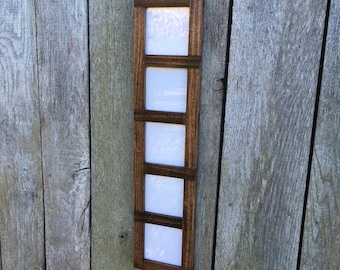 May include: A vertical wooden picture frame with four white square openings. The frame is made of dark brown wood and is mounted on a weathered wooden background. The frame is designed to hold four photos.