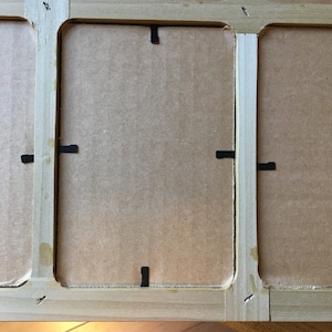 May include: A wooden picture frame with three openings, each with a cardboard backing and black paper tabs for holding the picture in place.
