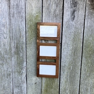May include: A vertical wooden picture frame with three rectangular openings. The frame is stained a dark brown color, and each opening has a white background. The frame is mounted on a weathered wooden surface.