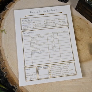 Może przedstawiać: Biała karta "Small Shop Ledger" ze złotym tekstem i obramowaniem, przeznaczona do gier fabularnych. Karta zawiera sekcje na nazwę sklepu, ekwipunek i rzadkie zapasy, z polami wyboru i listami przedmiotów. Kości i drewniane pudełko w tle.