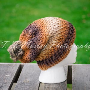 May include: A brown, beige, and black crocheted beanie with a fluffy pom pom. The beanie is on a mannequin head.