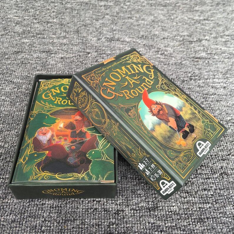 K&ouml;nnte beinhalten: Eine Kartenspielschachtel mit dem Titel Gnoming A Round, dunkelgr&uuml;nem Hintergrund und goldenen Verzierungen. Die Schachtel zeigt einen Gnom mit roter M&uuml;tze und kariertem Kilt. Die Schachtel ist ge&ouml;ffnet und zeigt ein Kartendeck mit Illustrationen.