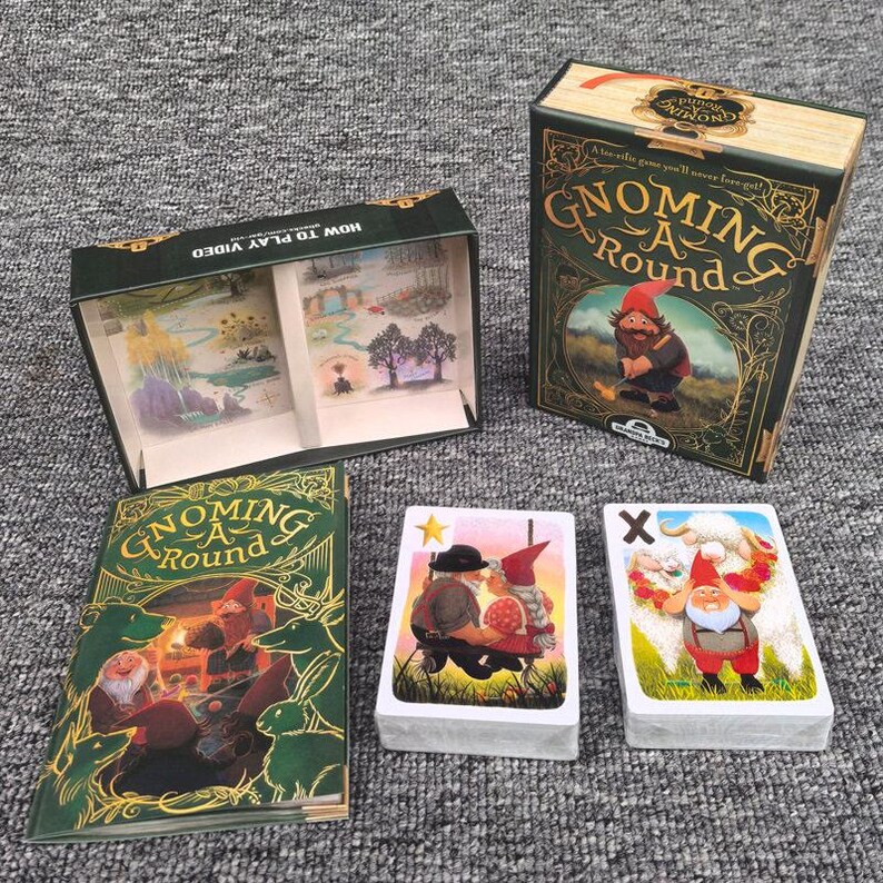 K&ouml;nnte beinhalten: Ein Gnoming A Round Kartenspiel-Set bestehend aus einer rechteckigen Schachtel mit Gnom-Illustration, einer offenen Aufbewahrungsbox mit Trennw&auml;nden, einem Regelheft mit gr&uuml;nem Einband und zwei Kartendecks mit Gnom-Motiven und Symbolen.