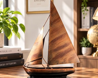 May include: A decorative metal sailboat sculpture featuring a weathered copper-toned finish on the sails and hull. The boat is mounted on a solid black rectangular base, showcasing a pointed mast with a small triangular flag at the very top.
