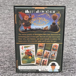 K&ouml;nnte beinhalten: Eine rechteckige Brettspielschachtel mit dem Titel Gnoming A Round, die eine Gnom-Illustration, einen goldenen Pokal und Spielkarten zeigt. Die Schachtel enth&auml;lt Spieldaten f&uuml;r 2-7 Spieler, Alter 7-99, und 15-30 Minuten Spieldauer, sowie beschreibenden Text und ein Foto von Grandpa Beck.